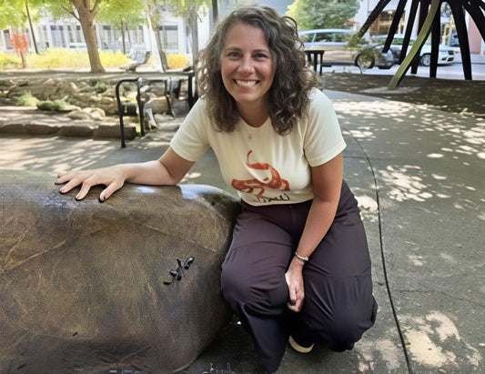 A smiling woman with curly hair kneels outdoors next to a large boulder with one of the Newts from Knoxville, wearing a cream graphic T-shirt and dark pants in a park-like setting.