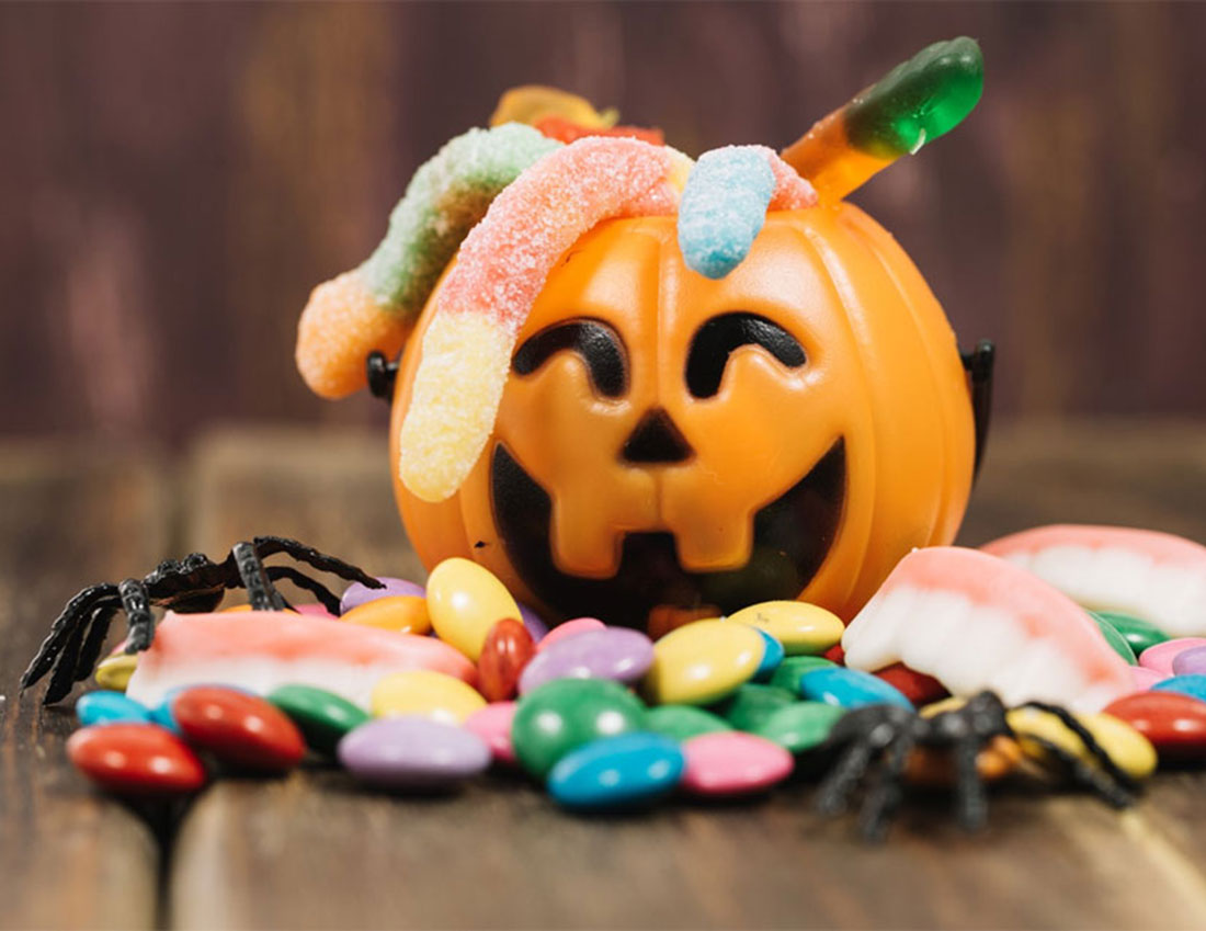 An orange jack-o'-lantern candy holder filled with colorful candies, topped with gummy worms, set on a wooden surface with plastic spiders around it.