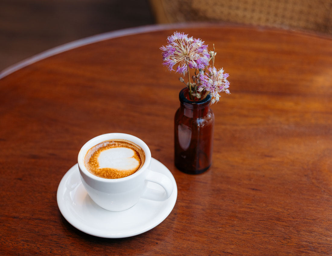 A latte in a white cup has a heart shape poured on top sitting on a medium brown colored round wooden  table sitting next to a small brown vase with purple flowers.