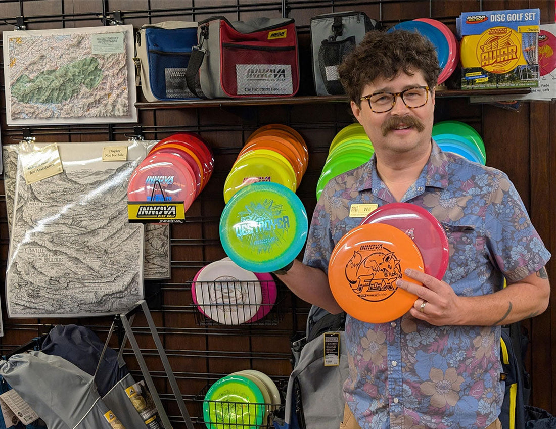 A man holding three disc golf discs standing in front of a disc golf display inside a store. Will, the outdoor/footwear manager at Mast Store Winston-Salem is an avid disc golfer.