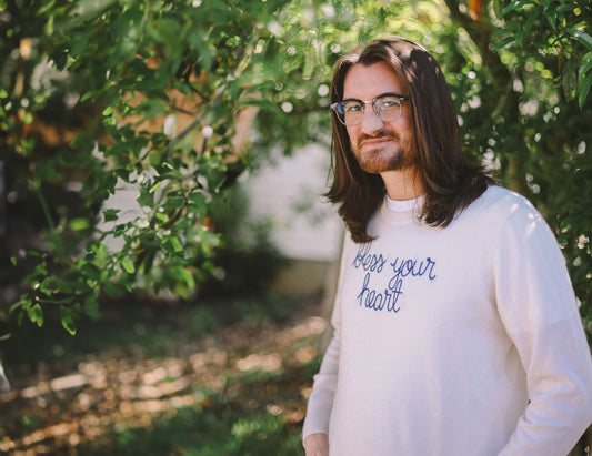 A male with long brown hair and glasses wears a long-sleeved shirt with the words Bless your Heart on it in a wooded setting.