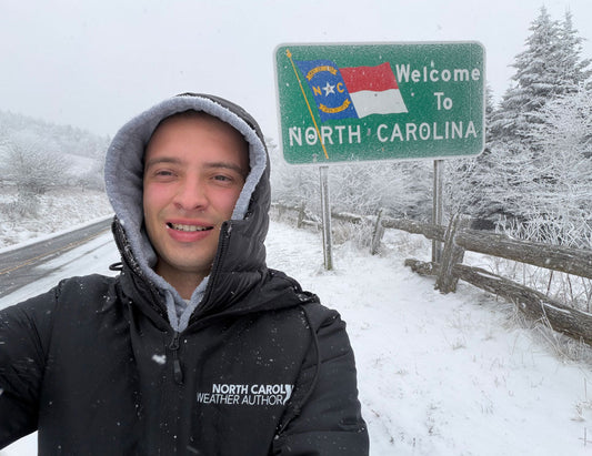 A man wearing a black winter jacket with a gray hooded lining takes a selfie next to a green 'Welcome to North Carolina' state sign in a snowy outdoor setting.