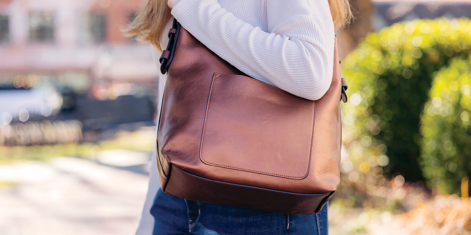 A person outdoors on a sunny day holding a Joy Susan brown leather bag, with a blurred background