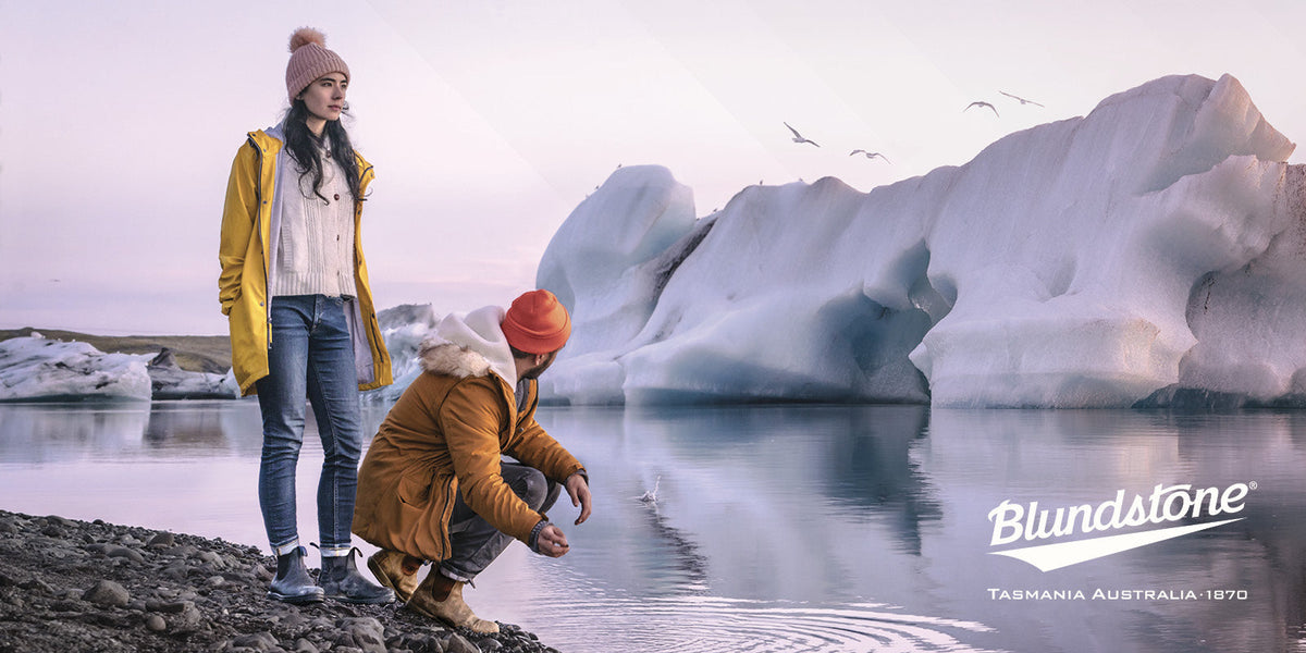 Two people in winter clothing and Blundstone boots stand by an icy lagoon surrounded by towering ice formations