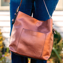 Person holding a brown Susan Joy leather handbag, with a blurred outdoor background.