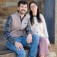 A man and a woman sitting together outdoors against a rustic stone wall. The man wears a beige vest over a blue plaid shirt and jeans, and the woman wears a cream knit cardigan, glasses, and mauve pants.