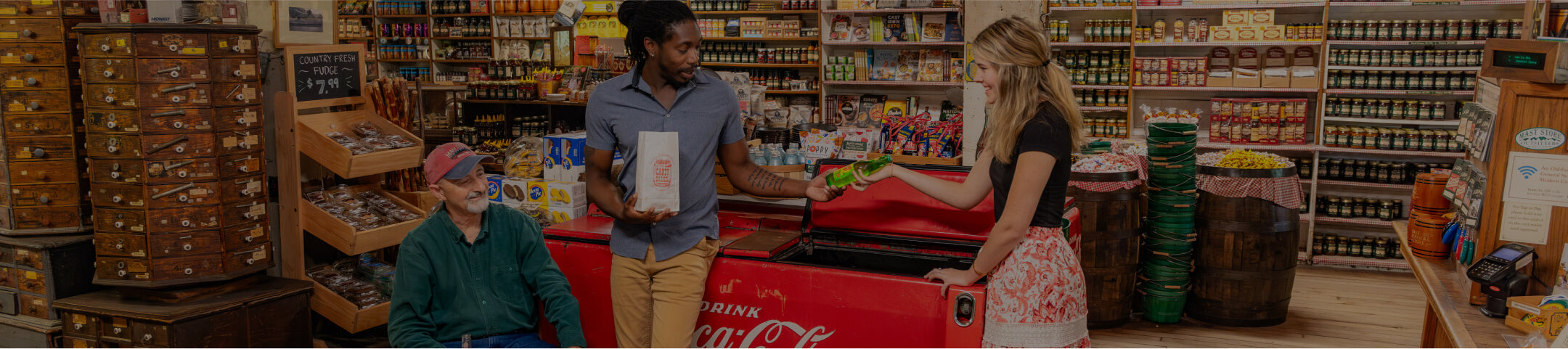 Three people hanging out by the Coca-Cola cooler. A lady is passing a glass bottle of soda to a gentleman holding a bag of candy. 