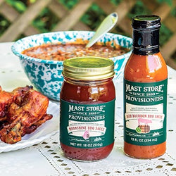 Two Mast Store sauce products displayed on a white tablecloth outdoors: a jar of Moonshine BBQ Sauce with a gold lid and a bottle of Beer Bourbon BBQ Sauce with a black cap, alongside a bowl of soup and cooked chicken.
