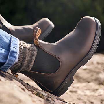 A close-up of a person wearing Blundstone brown leather boots and a knit sock, sitting outdoors on a rock with another boot blurred in the background.