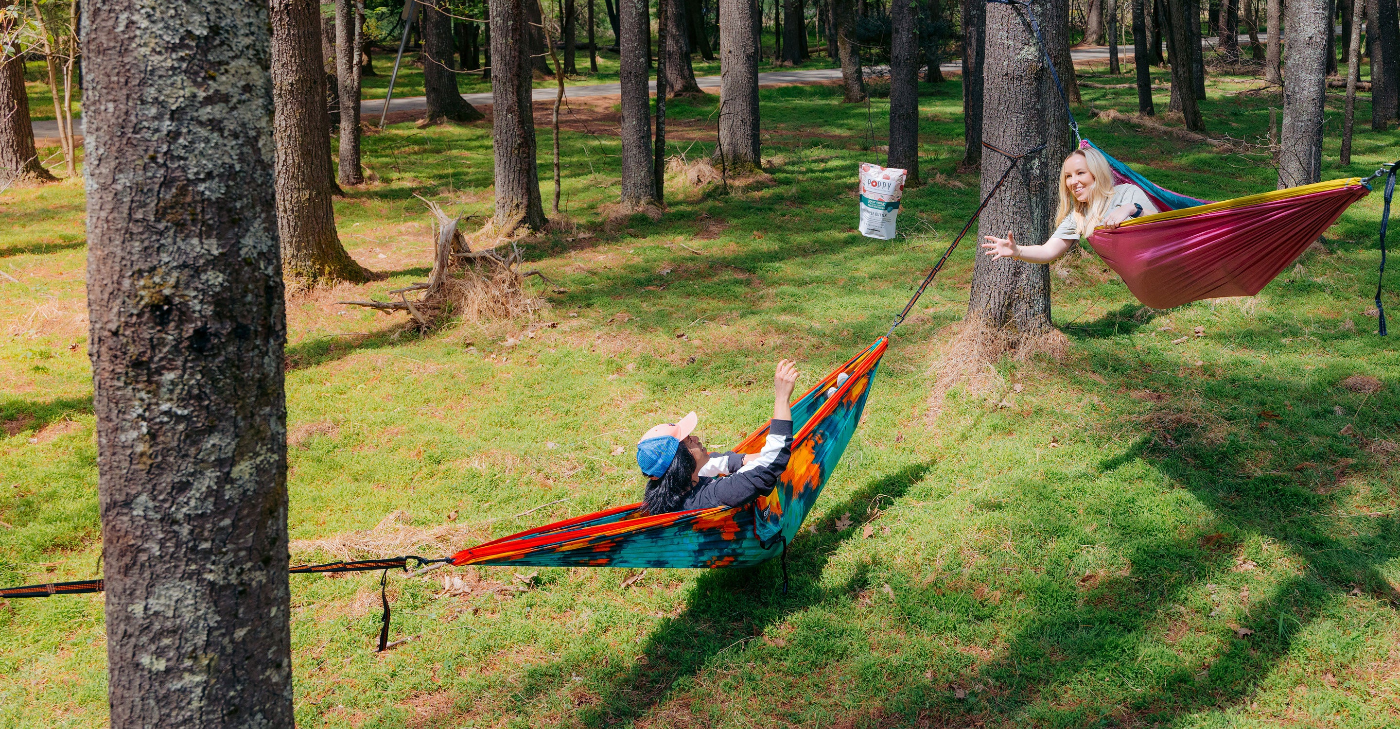 Two people lounging in bright ENO hammocks in the woods, laughing and sharing Poppy popcorn