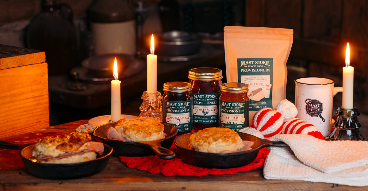 Cozy holiday breakfast scene featuring ham biscuits in cast iron skillets, alongside Mast Store Provisioners jams and biscuit mix, a Mast Store mug, and candlelit holiday warmth