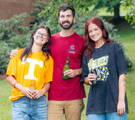 Three college students standing on a grassy lawn outdoors, wearing graphic T-shirts for the University of Tennessee, South Carolina Gamecocks, and Appalachian State Mountaineers